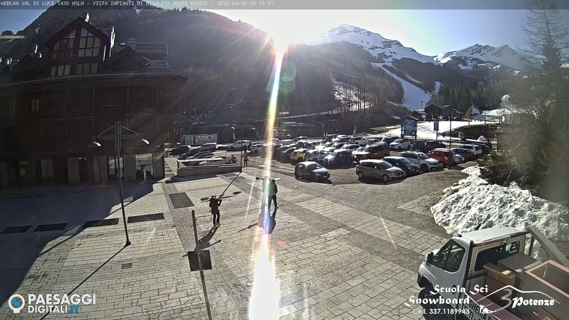 A sunny day in a mountain town shows a paved plaza with several people, a parking lot full of cars, and a building in the background, with snow-capped mountains and a ski slope visible in the distance.
