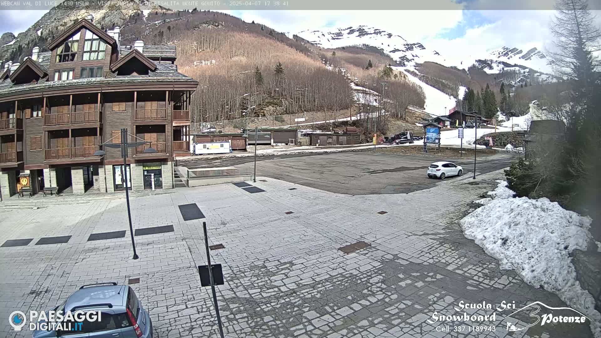 A paved plaza with a multi-story building and a few parked cars sits in front of a snow-capped mountain under a mostly sunny sky.
