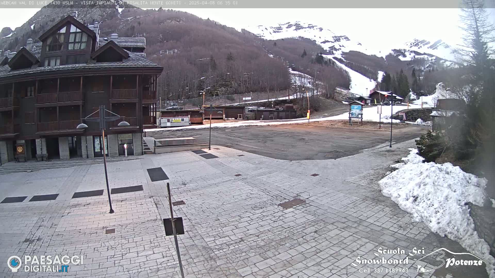A stone plaza sits in front of a multi-story building, with a snow-covered mountain and ski slope in the background under an overcast sky.