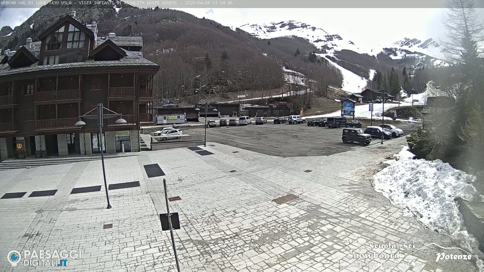 A paved plaza with a few cars parked in front of a multi-story building, overlooking a snow-capped mountain with ski slopes under a partly cloudy sky.