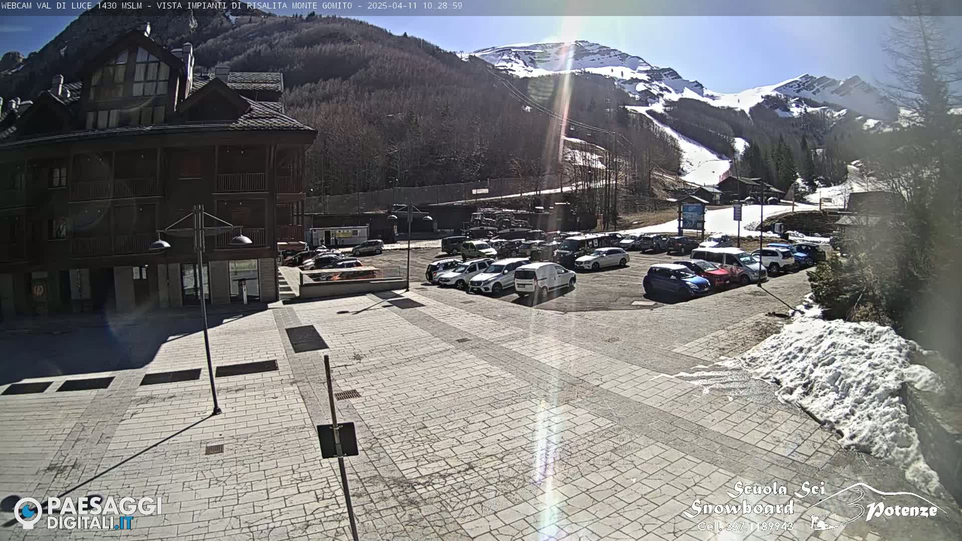 A sunny day reveals a parking lot full of cars in front of a building, with snow-covered mountains and ski lifts in the background.