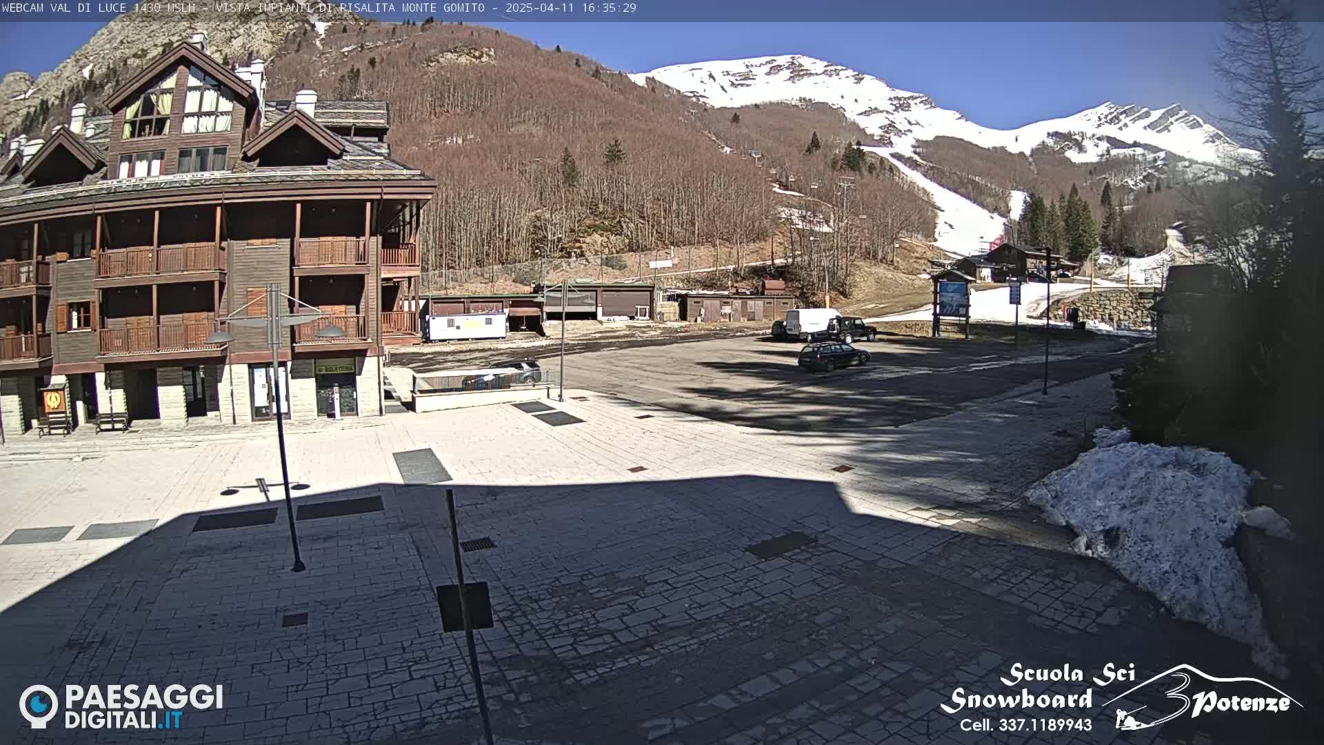 A sunny day shows a parking lot with several vehicles, a multi-story building, and snow-capped mountains in the background.