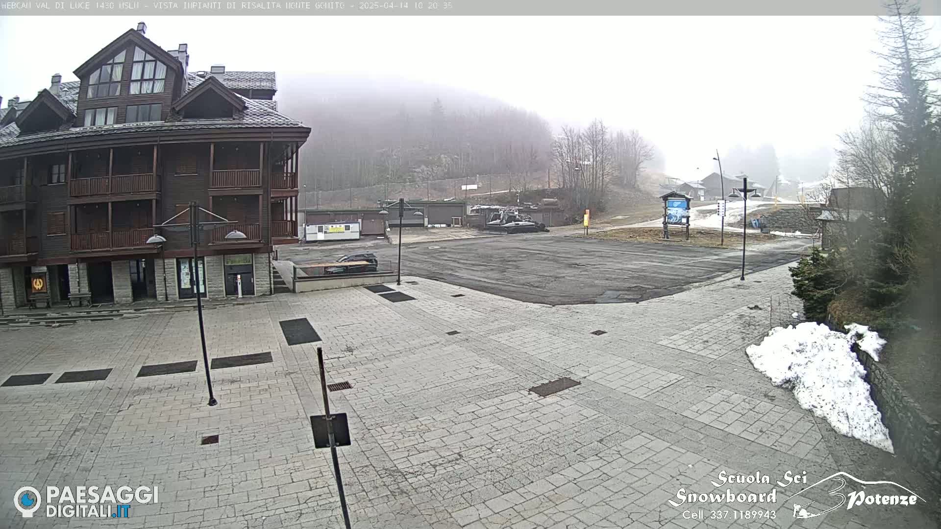 A foggy view of a paved plaza in front of a multi-story building, with a snowdrift and a mostly empty parking lot beyond.