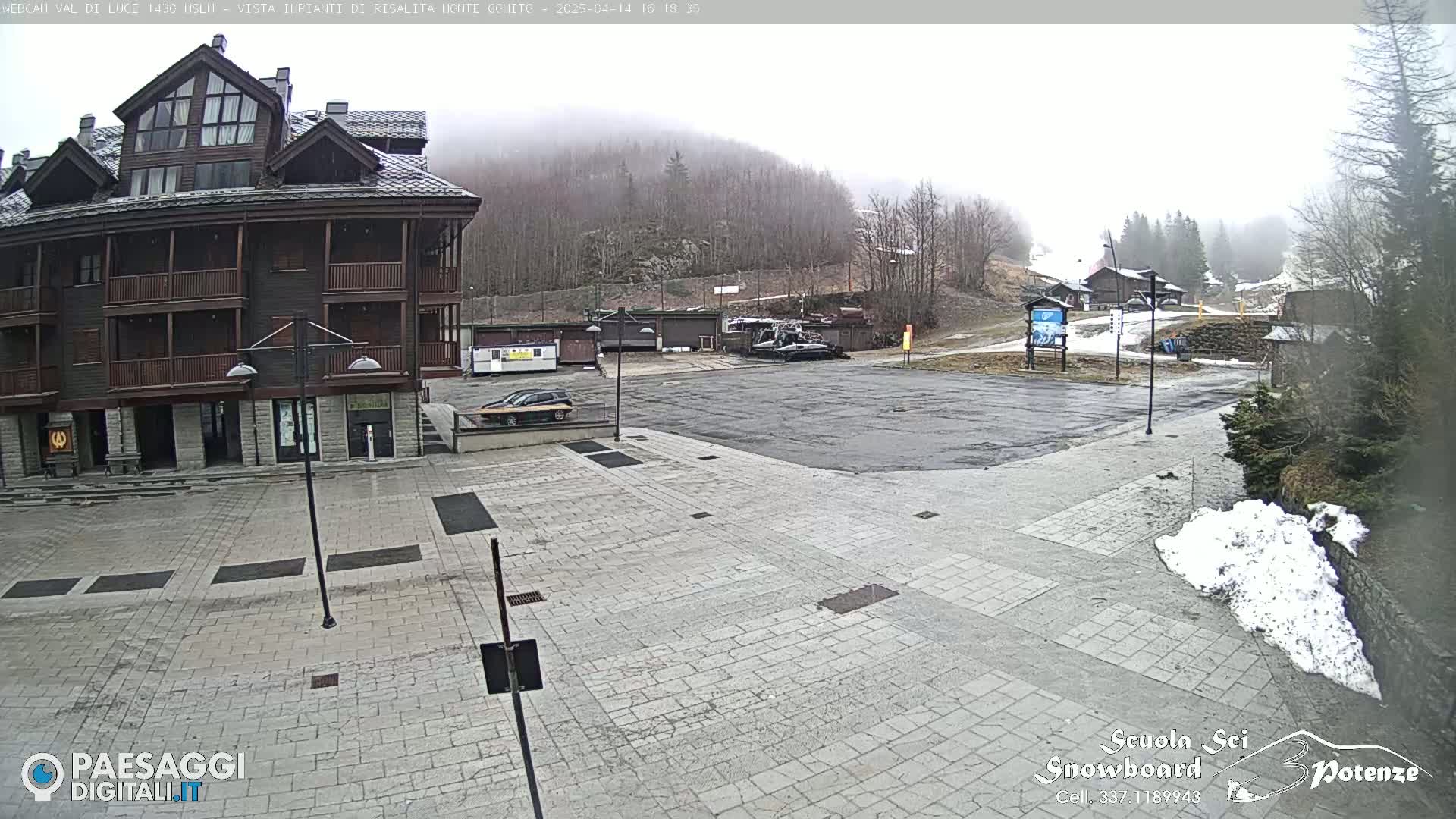 A paved plaza in front of a multi-story building is partially covered in snow under an overcast sky, with a mountainside visible in the background.