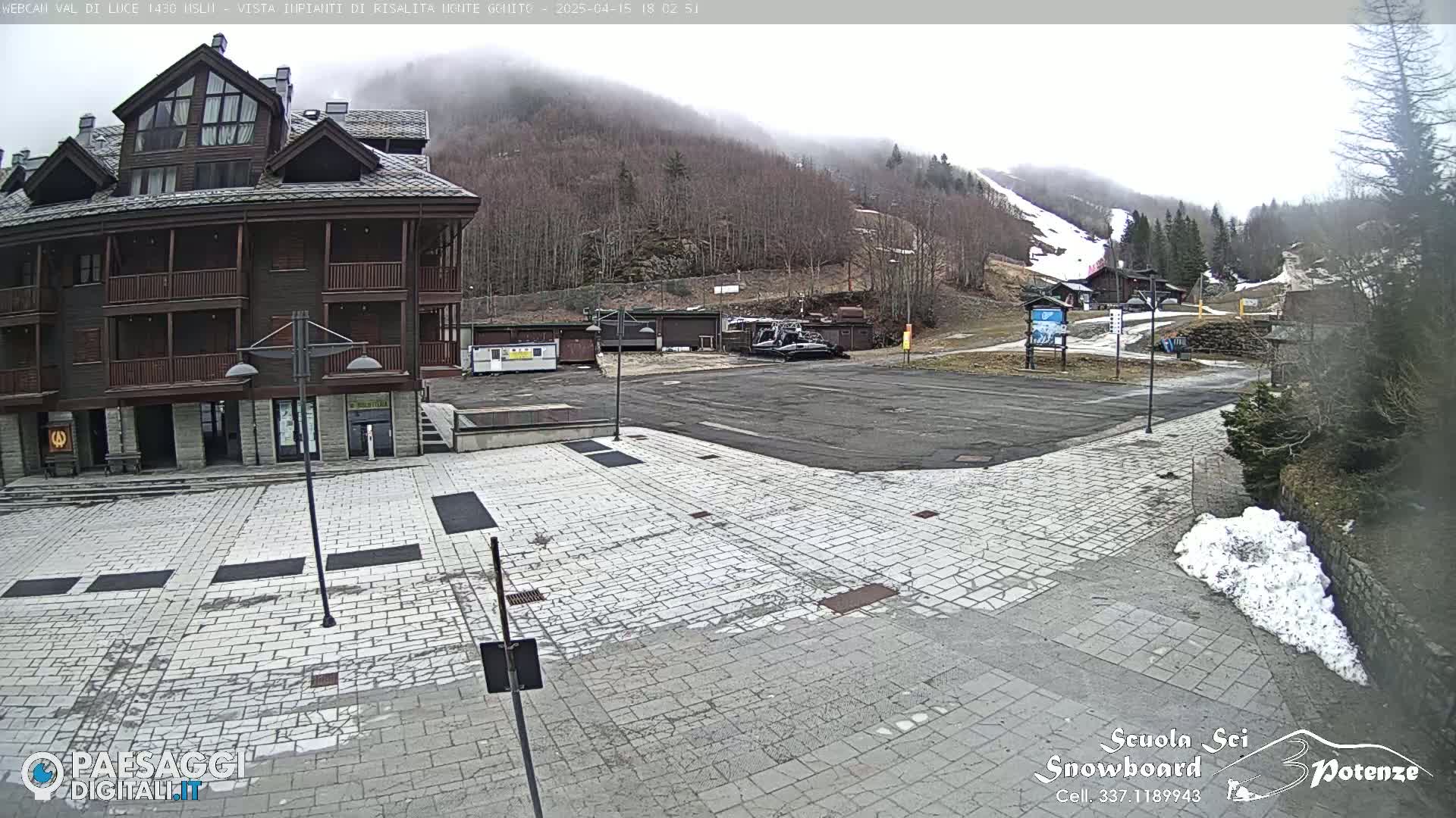 A stone plaza sits before a multi-story building, with a snow-covered mountain and ski slope visible in the background under an overcast sky.