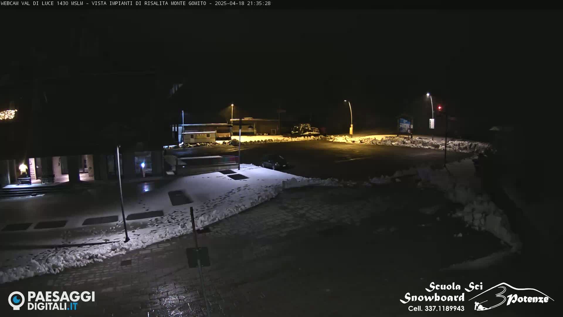 A nighttime view of a snow-covered parking lot and surrounding buildings, illuminated by streetlights.