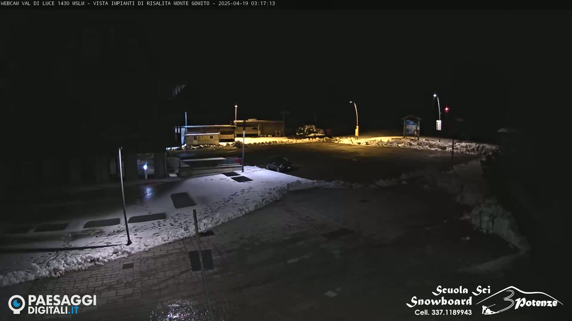A dark, nighttime scene shows a snow-covered parking lot with a few cars and buildings in the background.