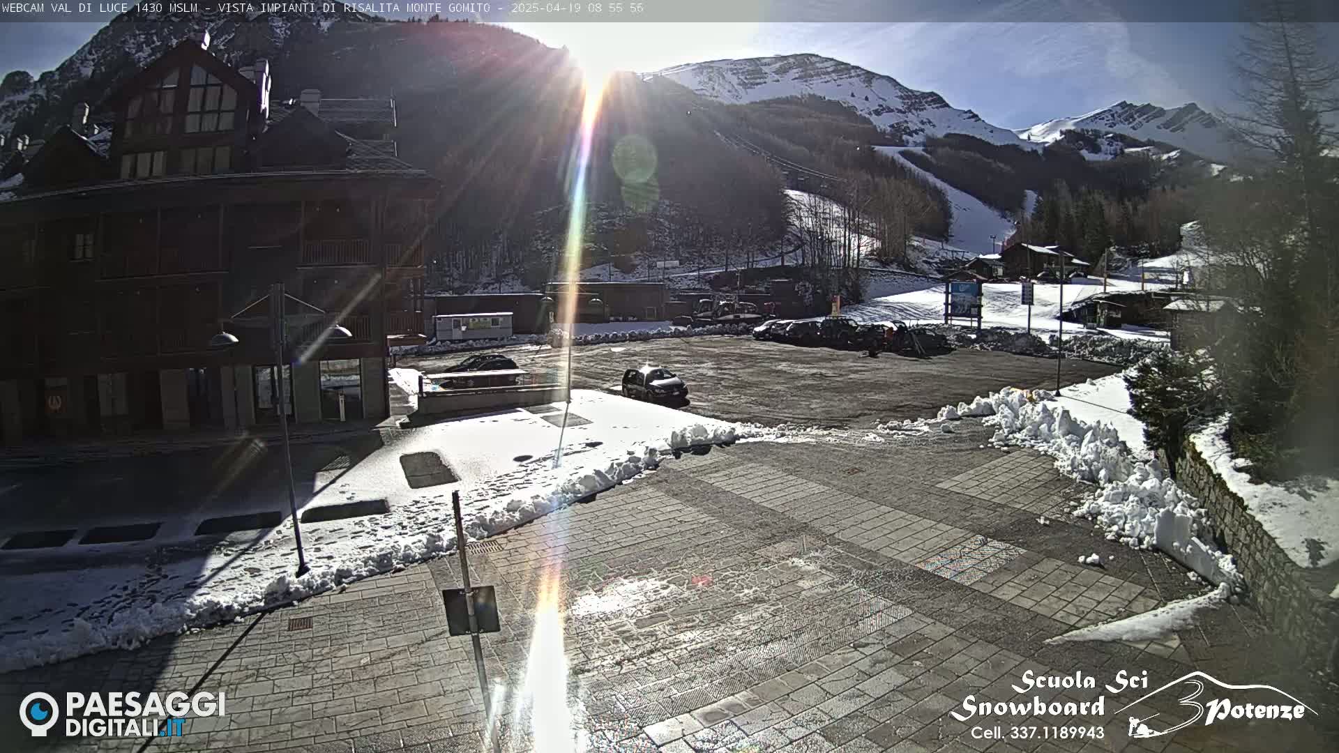 A sunny day in a snow-covered mountain town shows a parking lot with several cars, a multi-story building, and snow piles along a paved area.