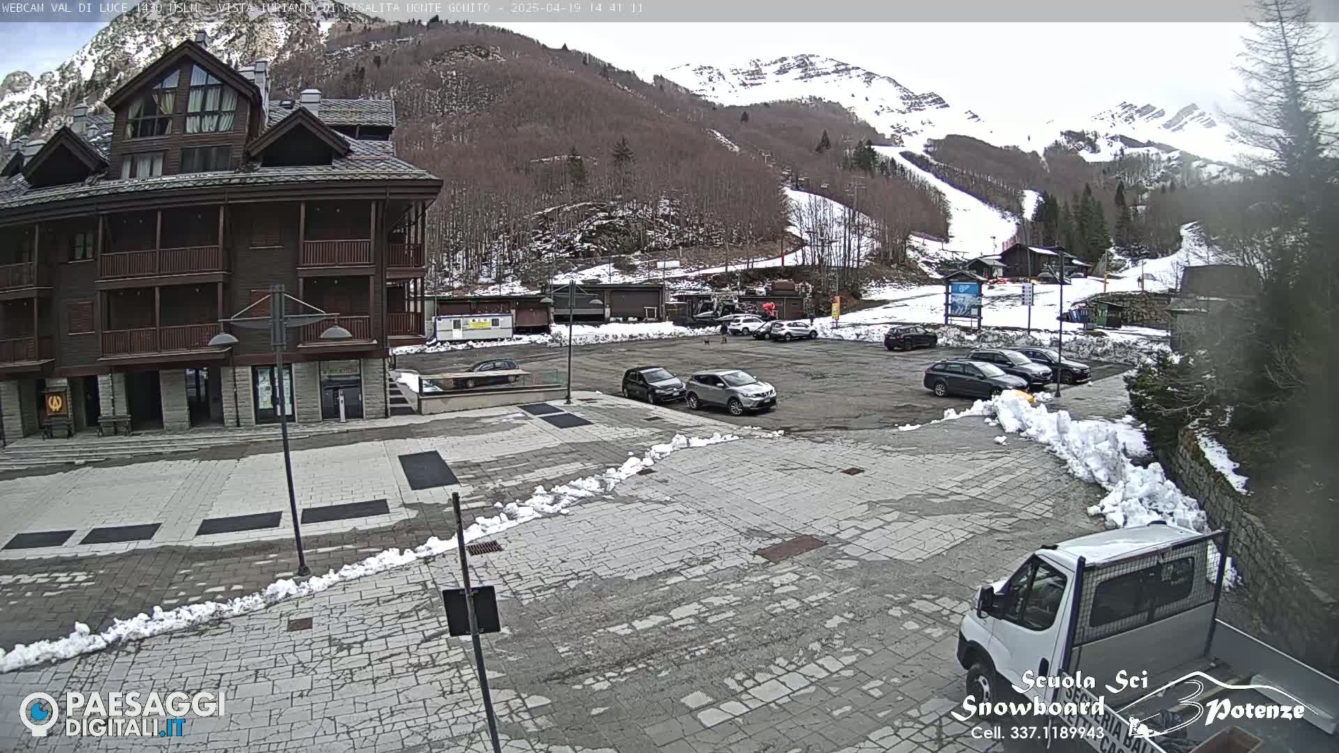 A paved parking lot with several cars and a van is in front of a multi-story brown building, with snow banks lining the edges and a snow-covered mountain in the background.