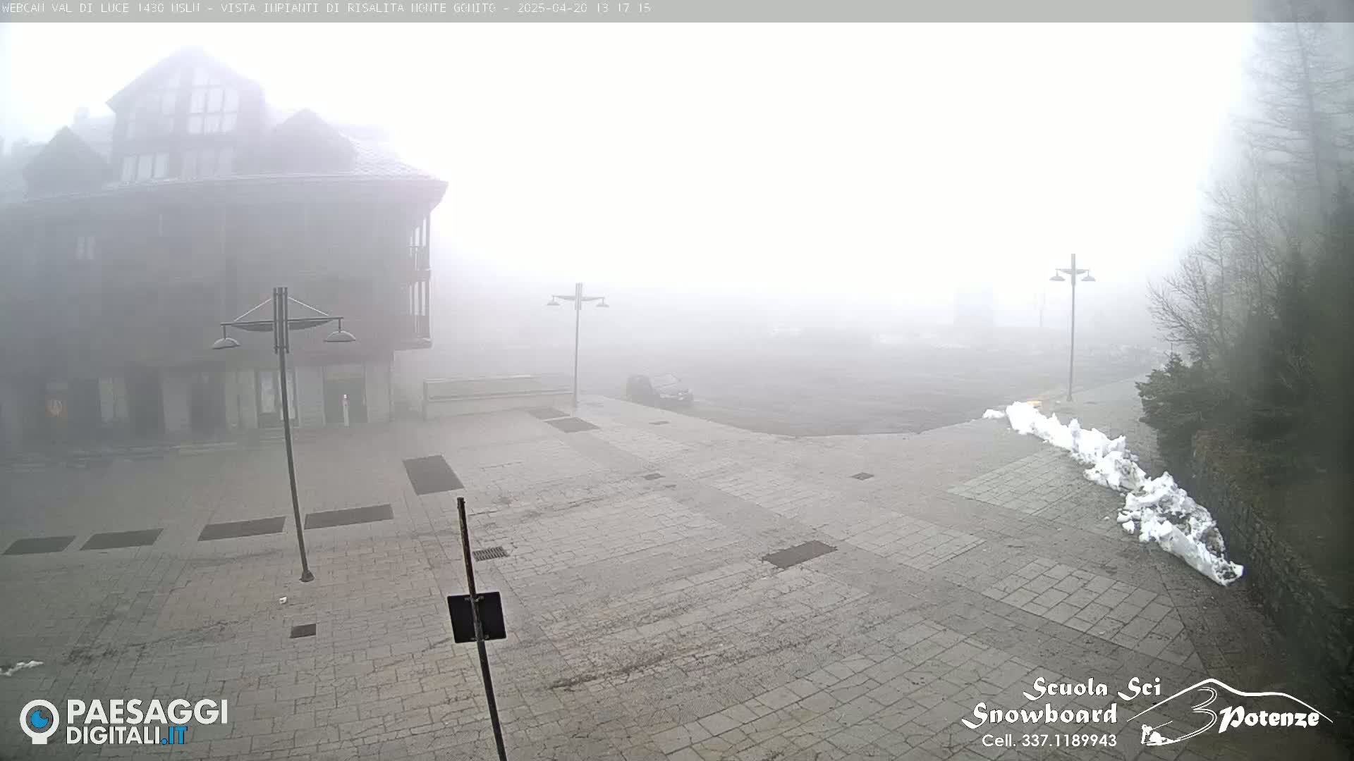 A stone plaza is shrouded in dense fog, with a multi-story building visible in the background and a small pile of snow in the foreground.