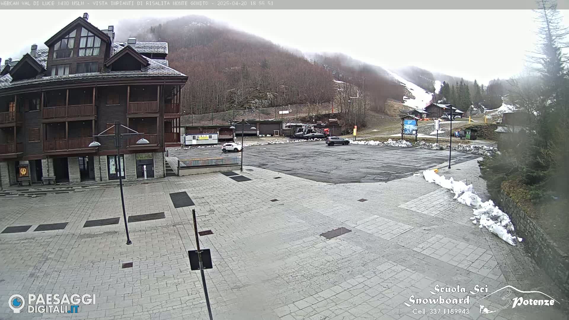 A multi-story brown building sits in a paved plaza with a few cars parked in a lot beyond it, overlooking a snow-covered ski slope on a cloudy day.