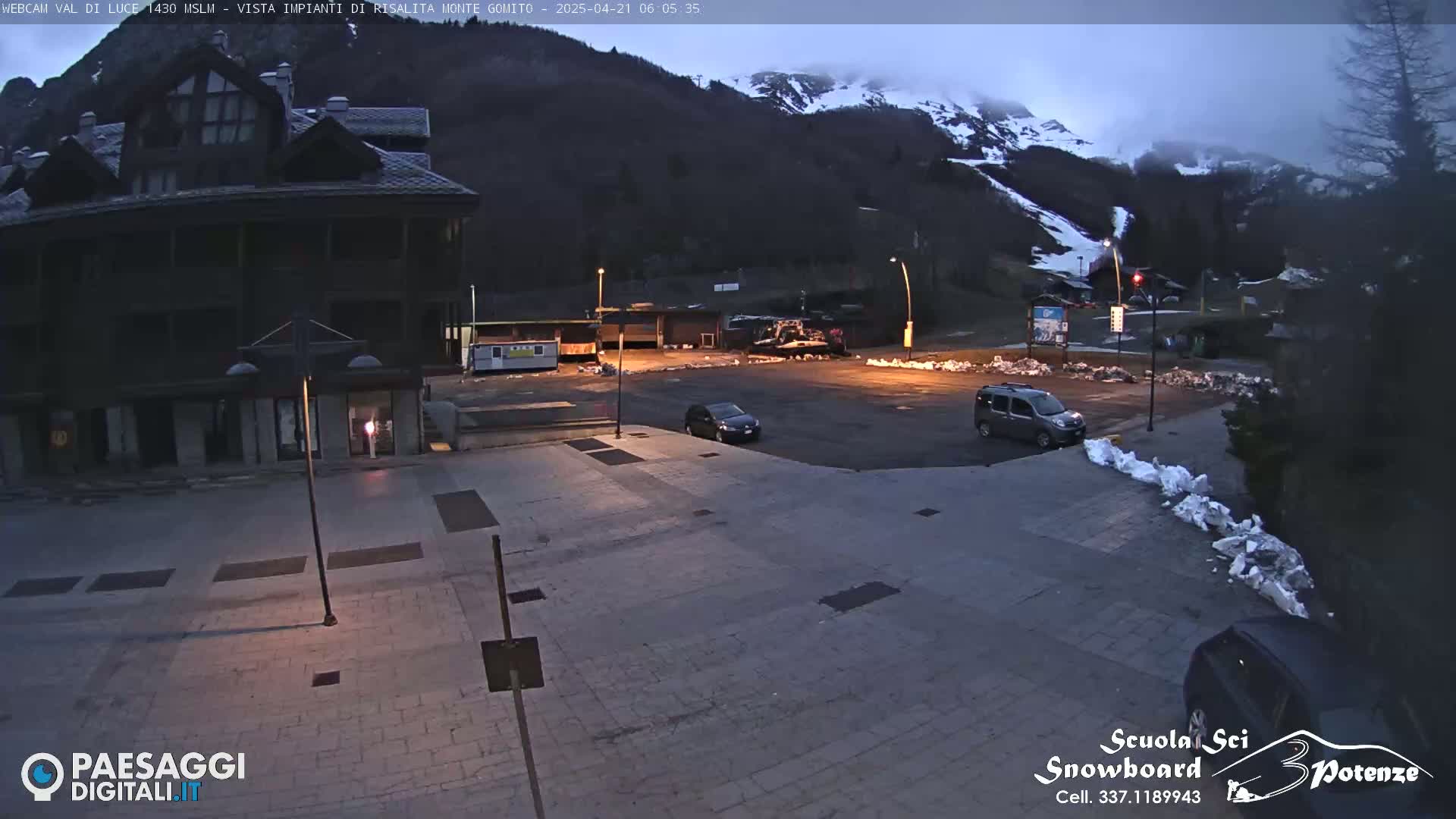 A mostly empty parking lot is situated in front of a multi-story building, with a snow-capped mountain in the background under a dark, pre-dawn sky.