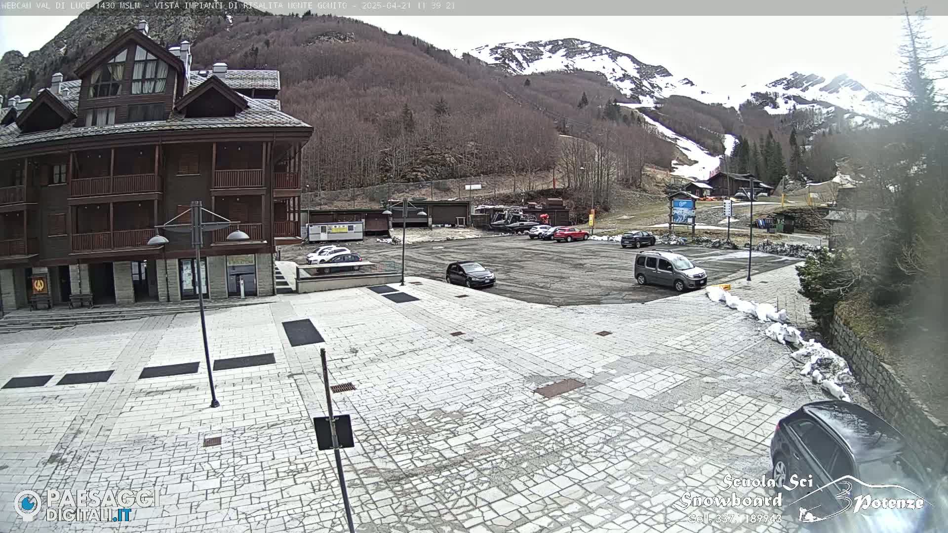 A stone plaza with parked cars in front of a multi-story brown building, overlooking a mountain with patches of snow under an overcast sky.