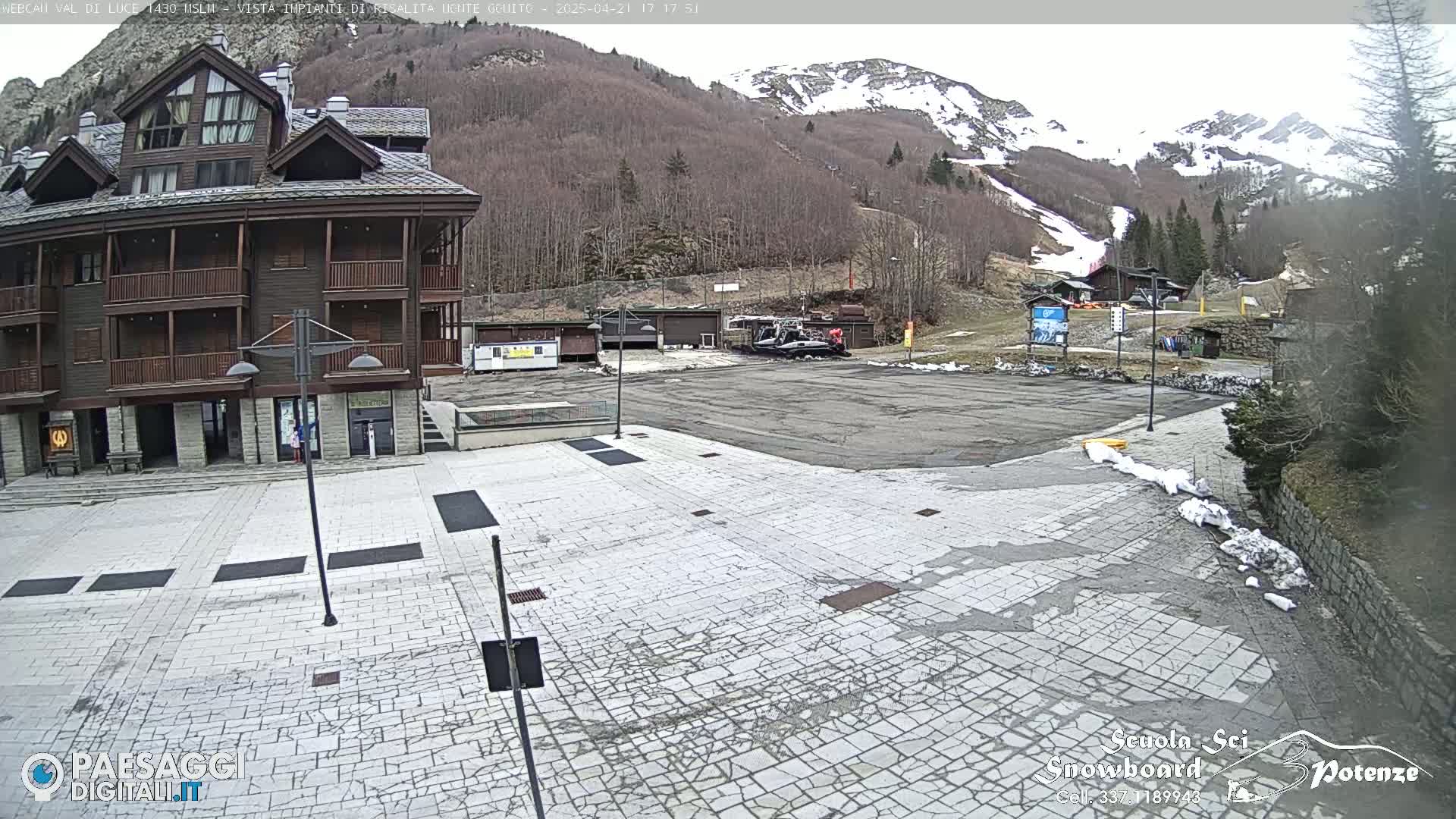 A stone plaza sits in front of a multi-story wood building, with a snow-dusted mountain and ski resort in the background under a cloudy sky.