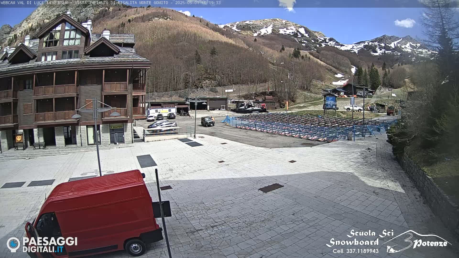 A red van is parked in a paved plaza in front of a multi-story brown building, with a mountainside visible in the background under a partly sunny sky.