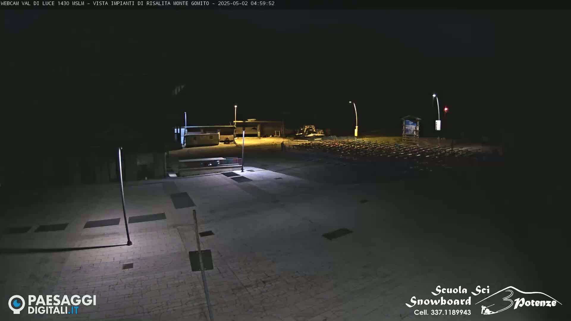 A nighttime, outdoor view of an empty plaza with several buildings and a large parking lot in the background.