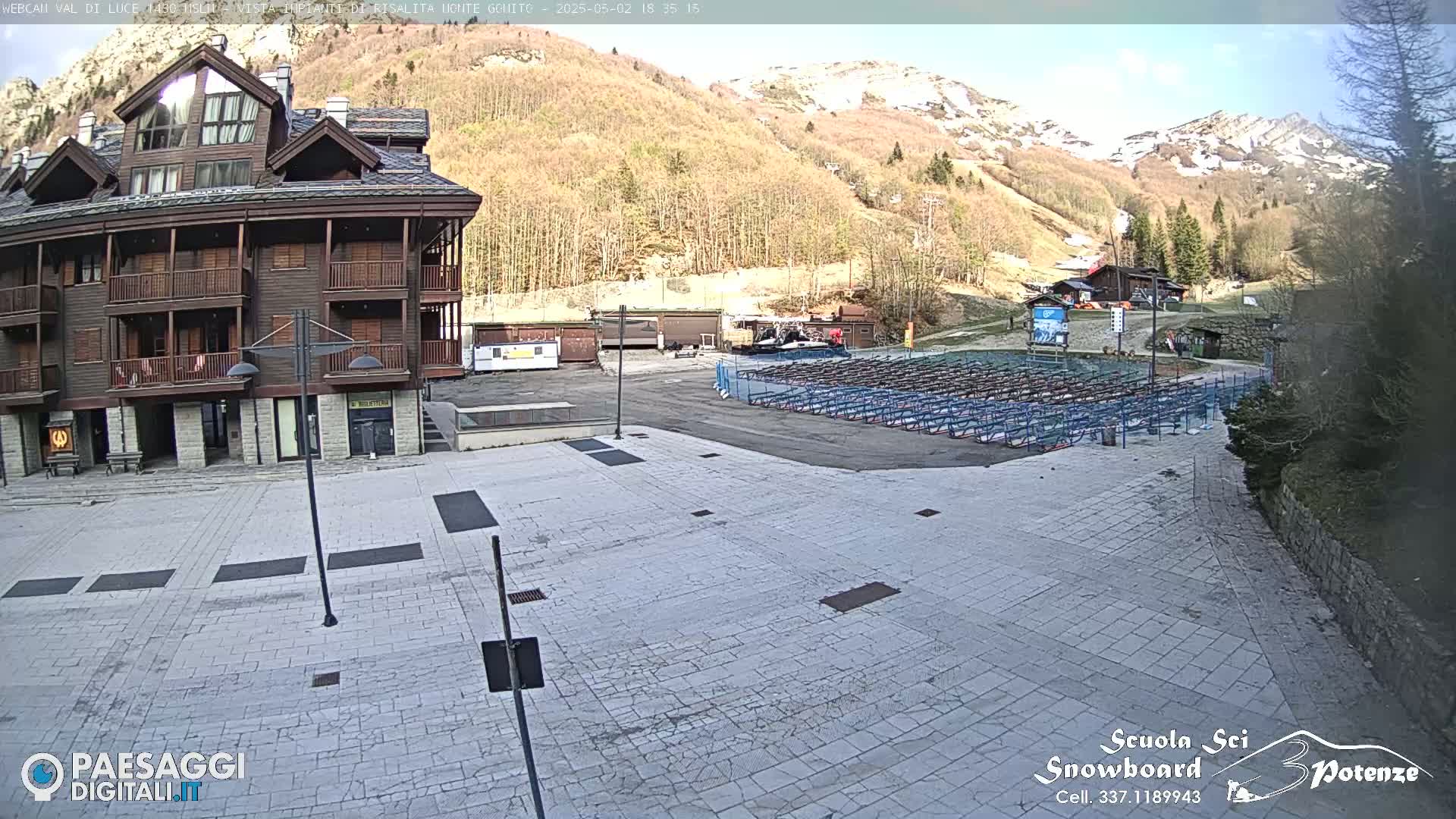 A large paved plaza sits in front of a brown multi-story building, with a snow-dusted mountain and sparsely leaved trees in the background under a mostly sunny sky.