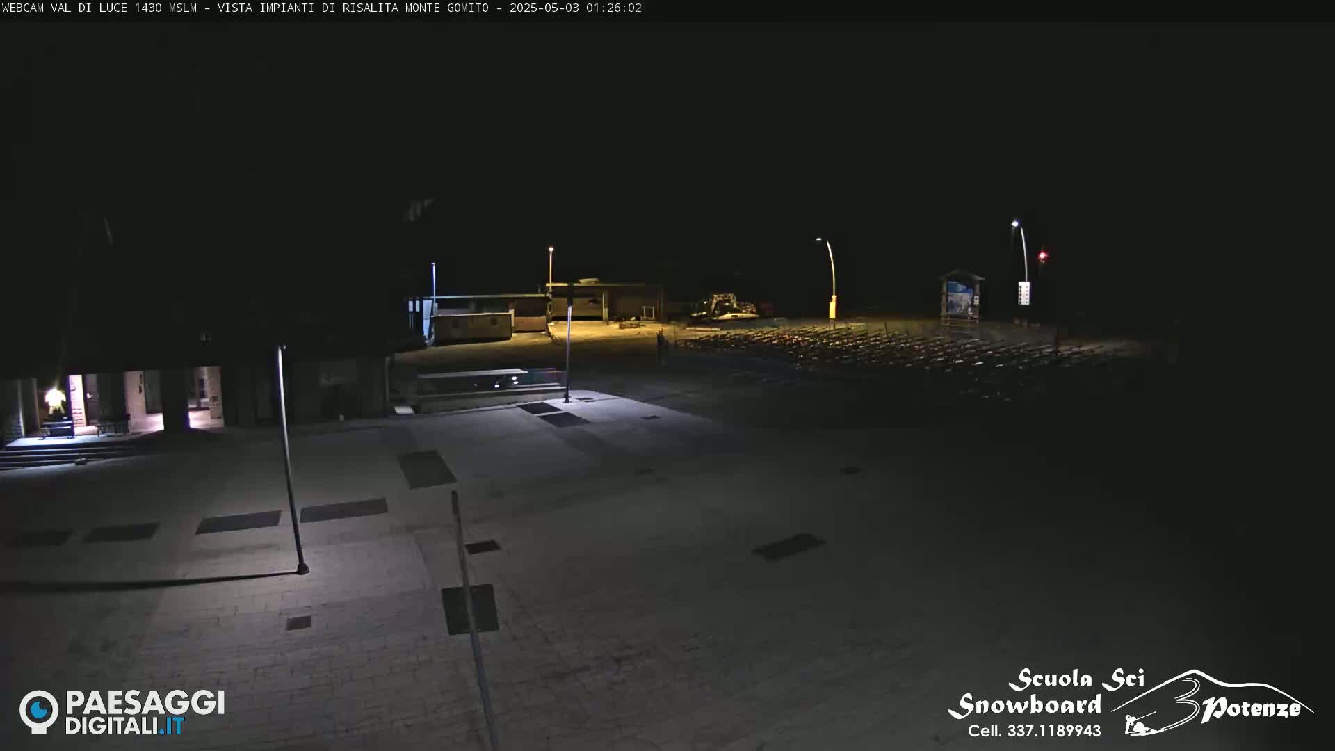 A nighttime, outdoor view of a mostly empty plaza with some buildings and a large area of what appears to be folded chairs or similar objects.