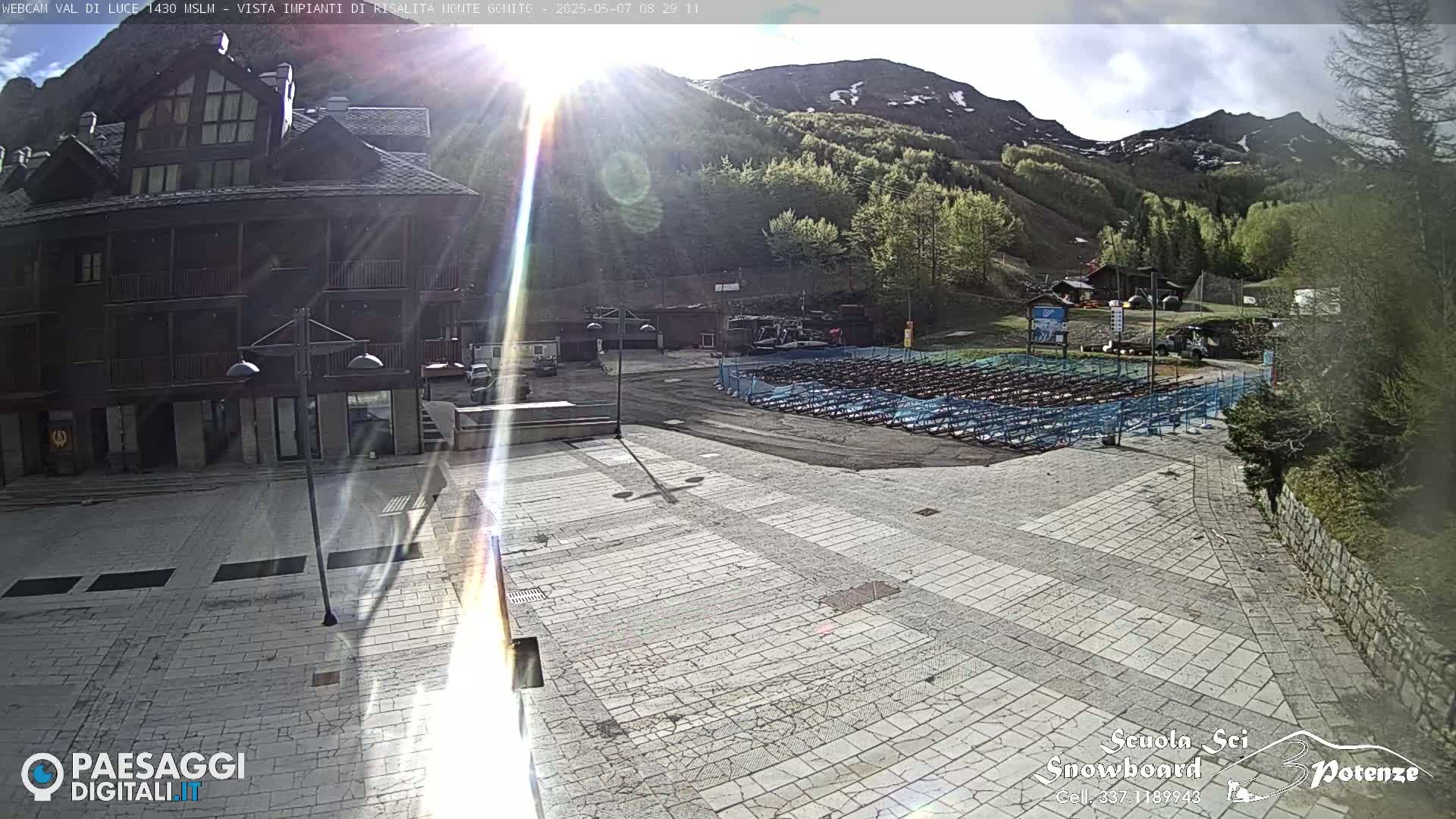A sunny day in a mountain town shows a paved plaza in front of a multi-story building, with rows of folded chairs in a fenced area beyond.