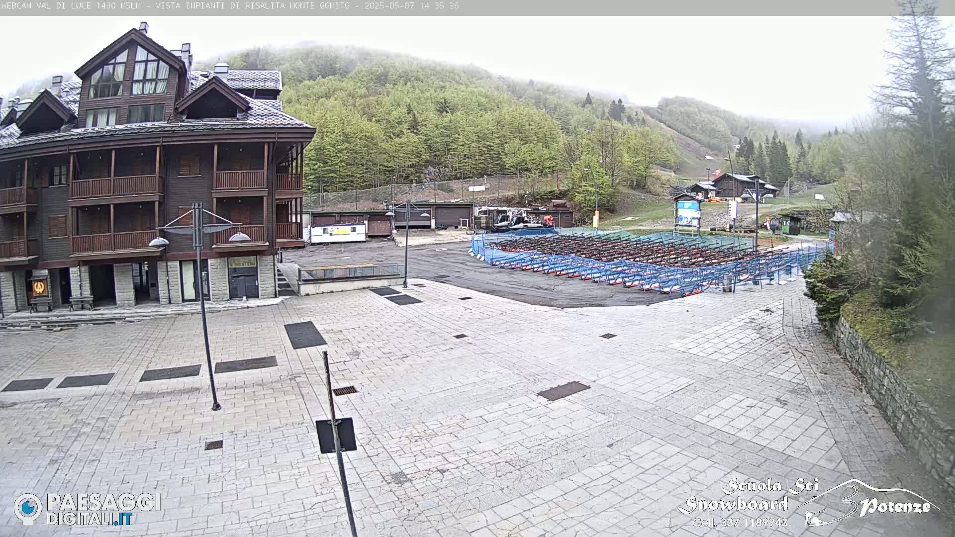 A multi-story building stands in a paved plaza overlooking a misty mountainside with numerous rows of what appear to be snowboard or ski racks.