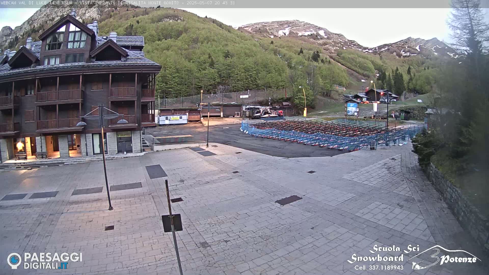 A multi-story building sits in a paved plaza overlooking a mountainside with green trees and snow-capped peaks under a mostly clear sky.