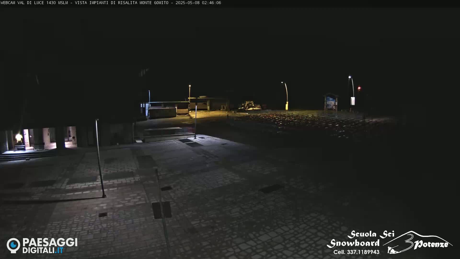 A dark, nighttime view of an empty paved plaza with some buildings in the background, illuminated by several streetlights.
