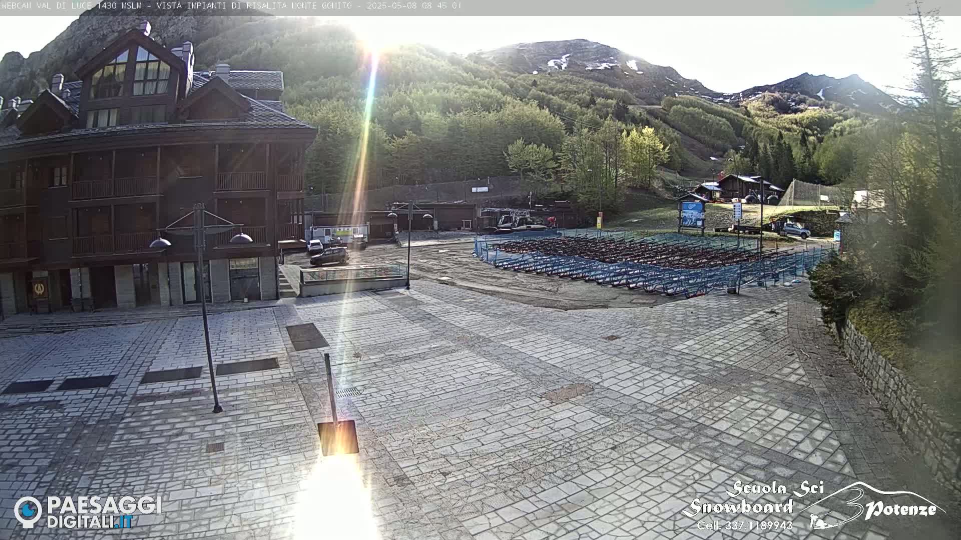 A sunny view of a stone plaza with a multi-story wooden building, a large area of stacked snowboards, and a mountainside in the background.