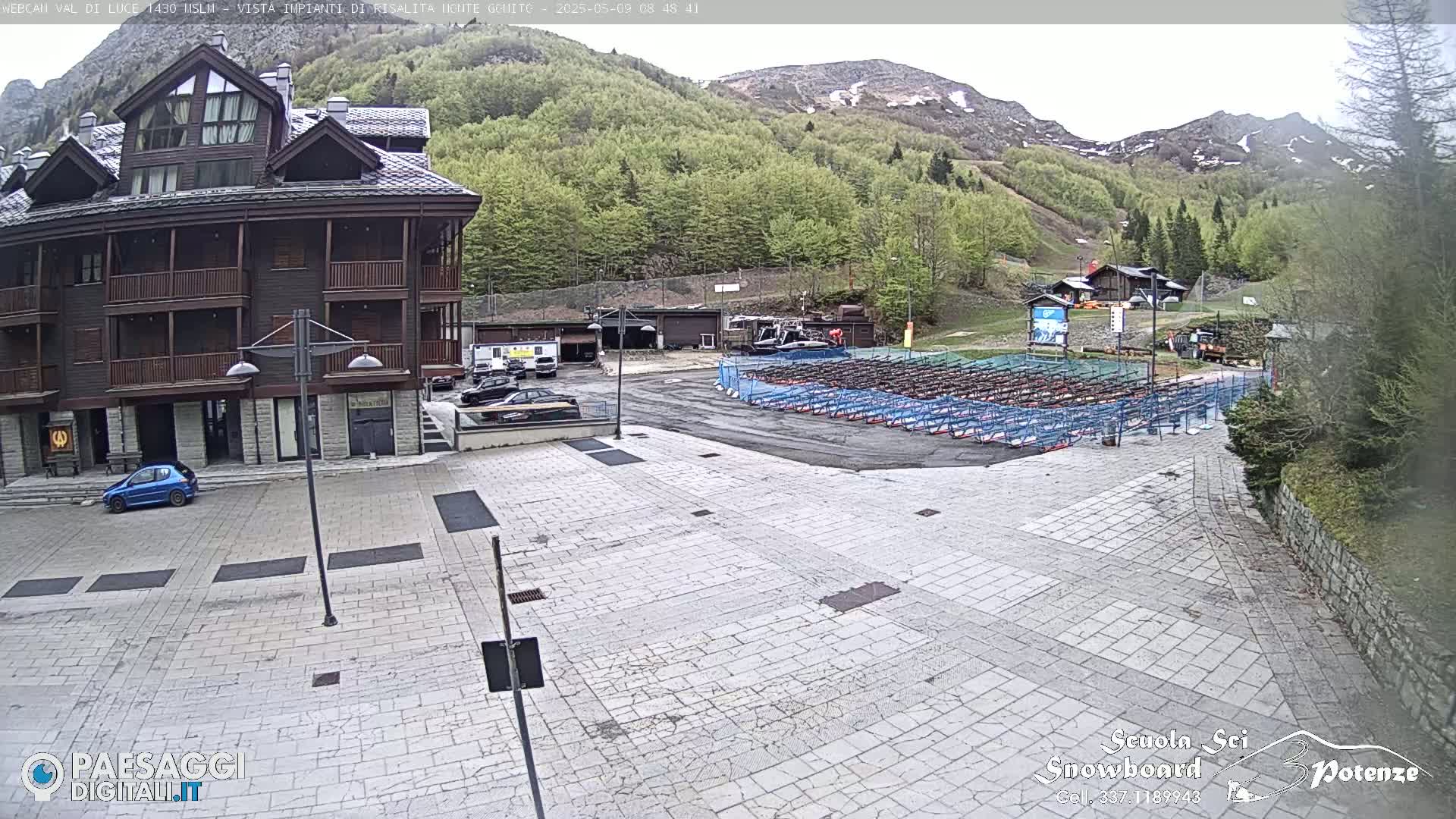 A paved plaza with a multi-story building, parked cars, and rows of what appear to be snow sport equipment racks is seen;  mountains and green trees are in the background under a partly cloudy sky.