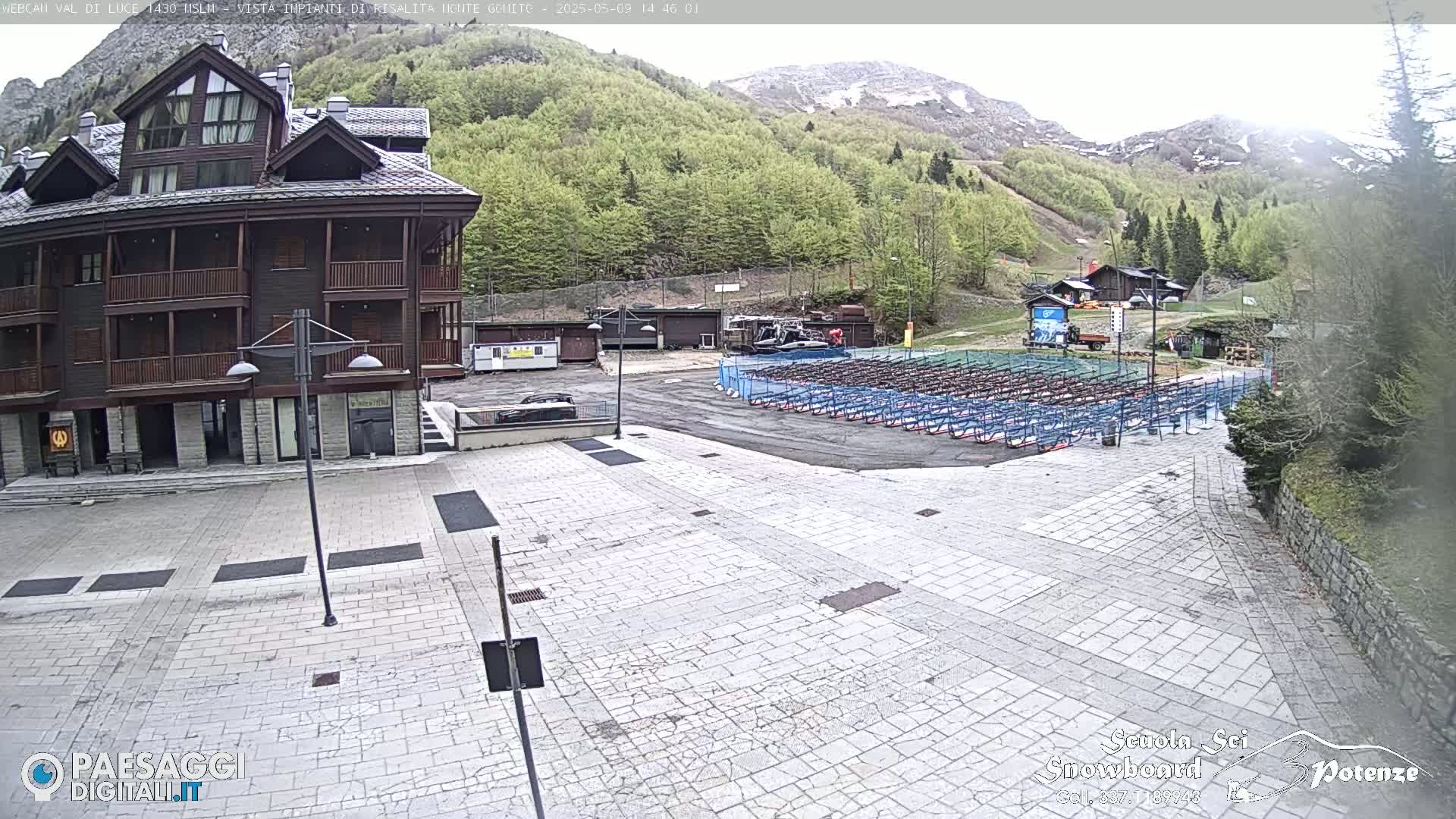 A paved plaza sits before a multi-story building, with rows of empty equipment racks in the background, nestled amidst a mountainside of lush green trees under an overcast sky.