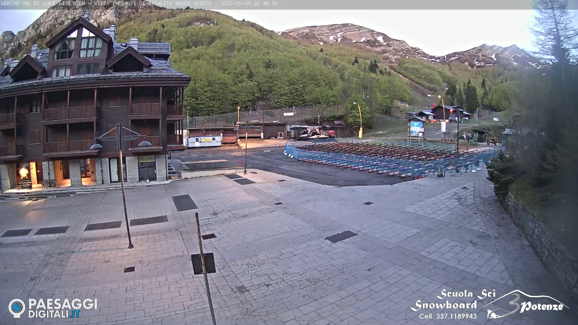 A paved plaza sits in front of a multi-story brown building, with numerous snowboards racked in the background against a backdrop of mountains under a partly cloudy sky.