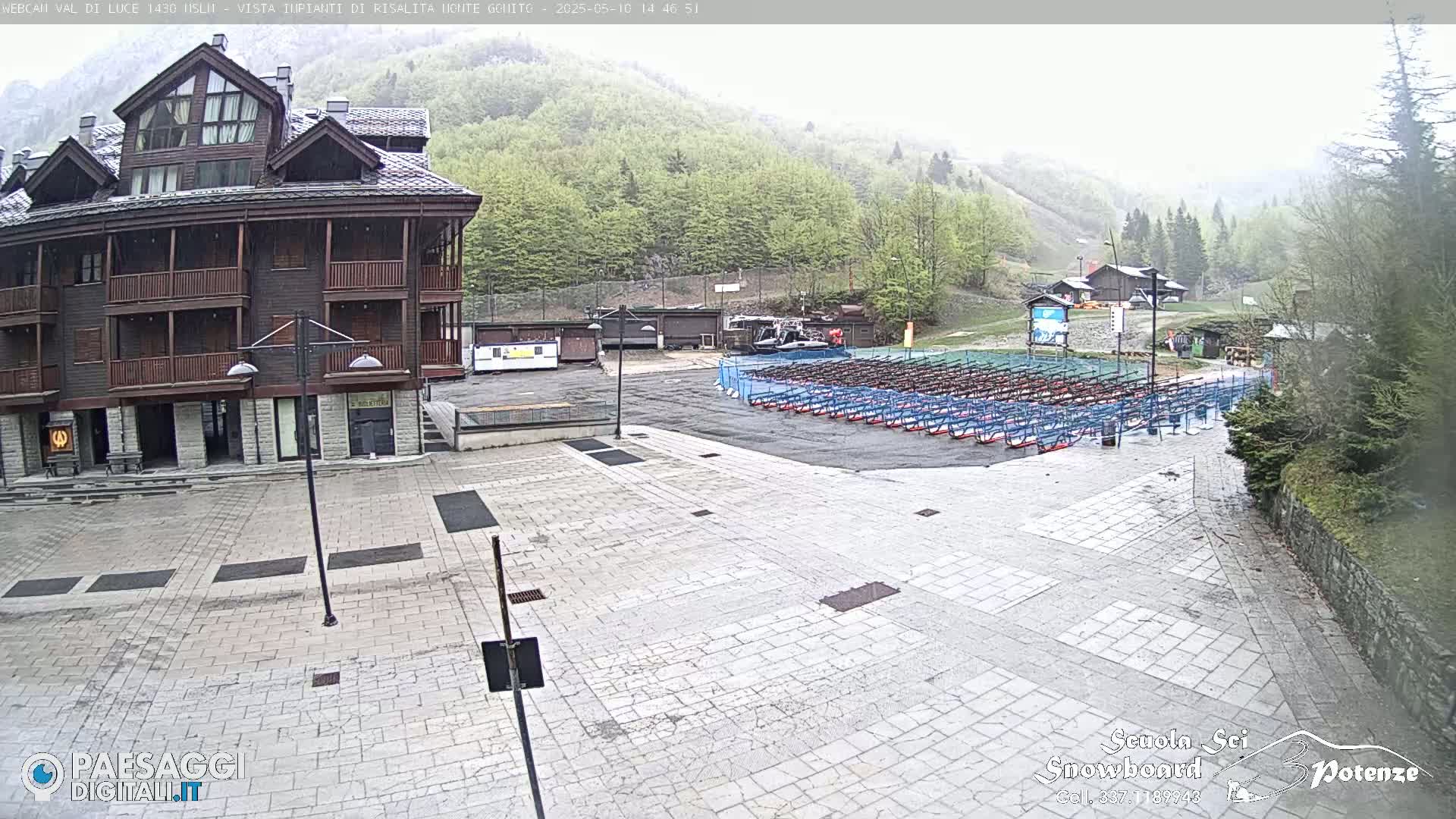 A multi-story building is situated in a paved plaza with rows of what appear to be ski or snowboard racks, under an overcast sky, lightly raining.