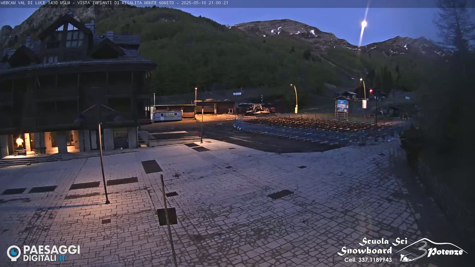 A nighttime view of a stone plaza next to a building, with rows of folded chairs in the background, and mountains under a twilight sky.