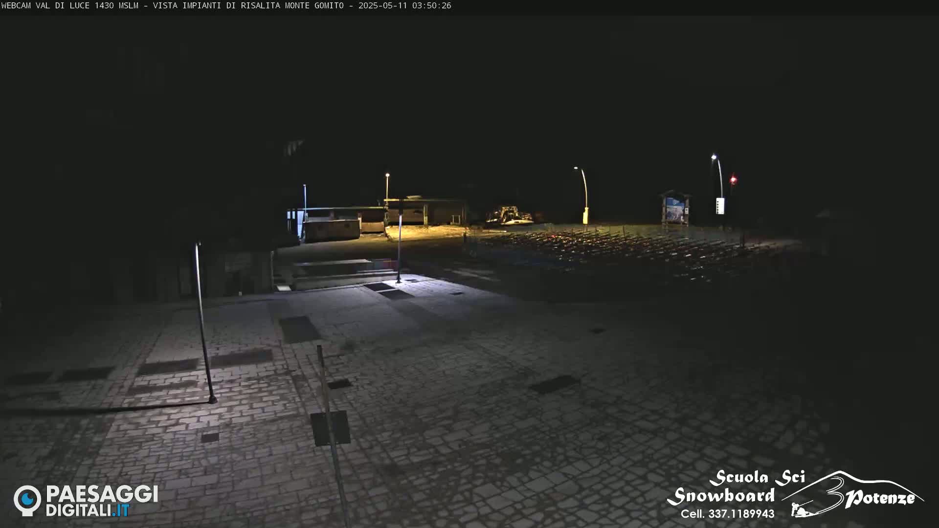 A dark, paved area at night is illuminated by several lights, showing a mostly empty parking lot with some structures in the background.