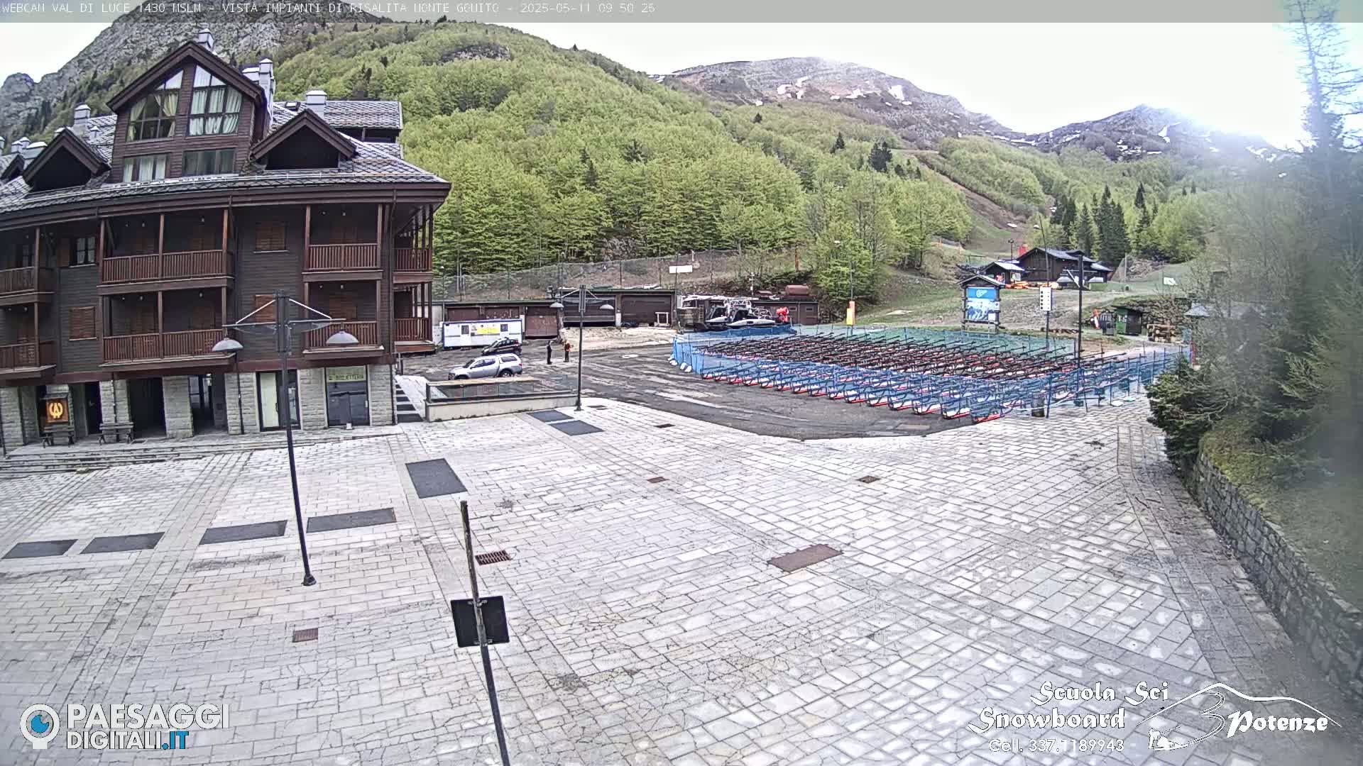 A stone plaza sits in front of a multi-story brown building, with rows of what appear to be ski or snowboard racks in the background, all set against a backdrop of green hills and mountains under an overcast sky.