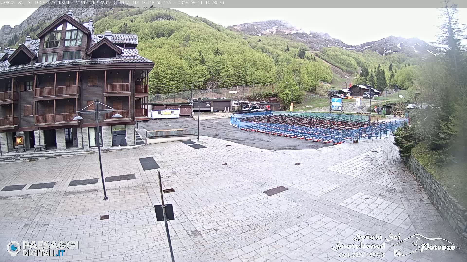 A stone plaza with rows of ski racks sits in front of a multi-story building, overlooking a mountainside with green trees and some patches of snow under an overcast sky.