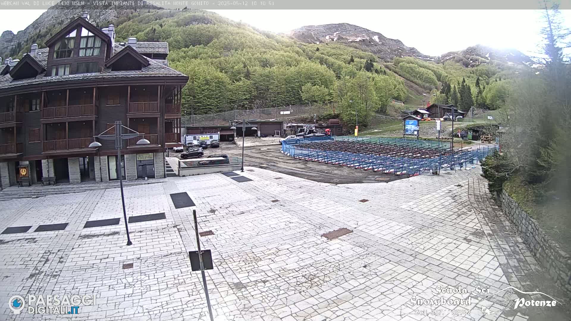 A mostly empty paved plaza sits in front of a multi-story building, with a mountainside covered in lush green trees in the background under a partly cloudy sky.