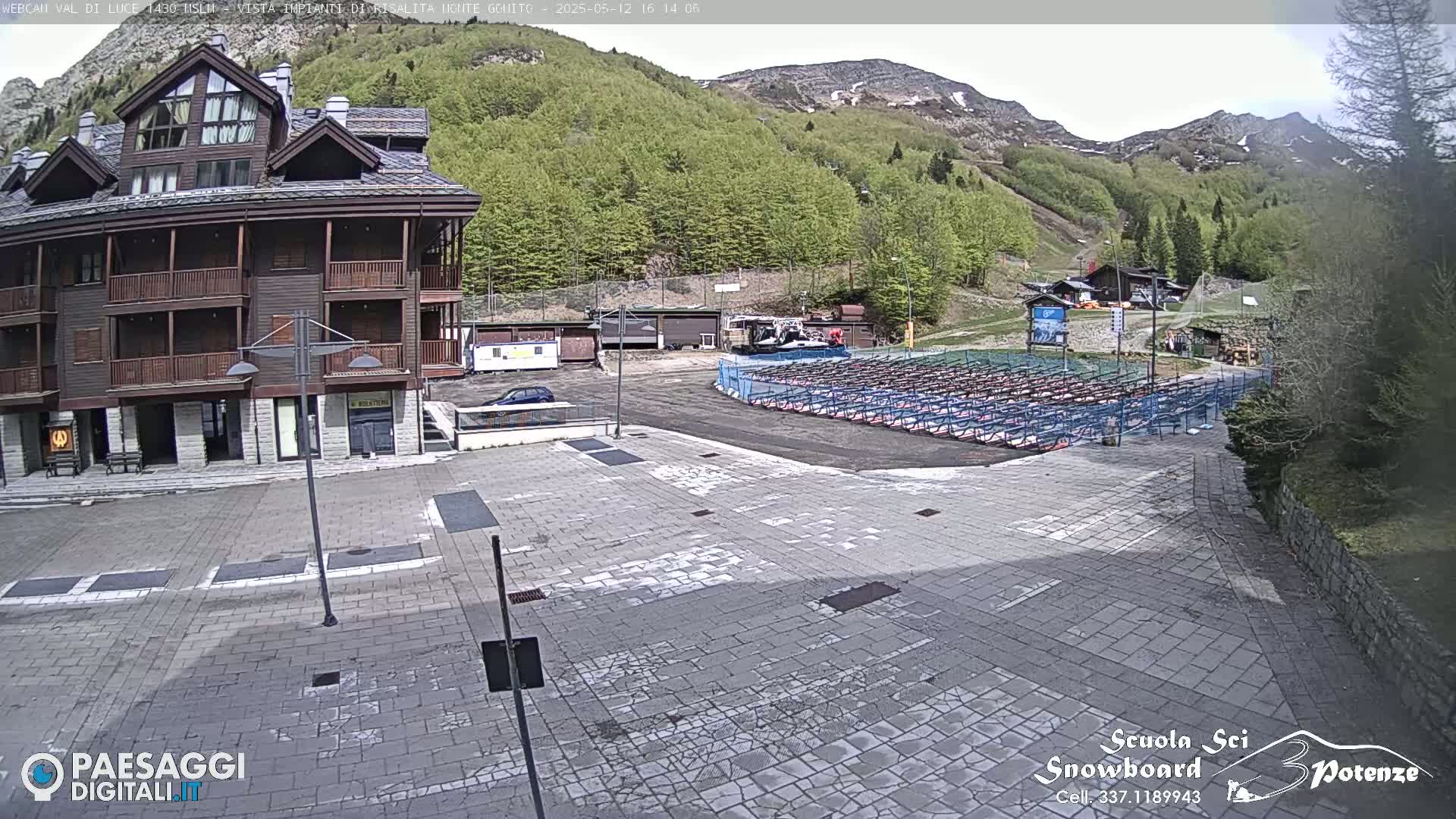 A paved plaza sits in front of a multi-story building, with a mountainside covered in lush green trees in the background under a mostly clear sky.