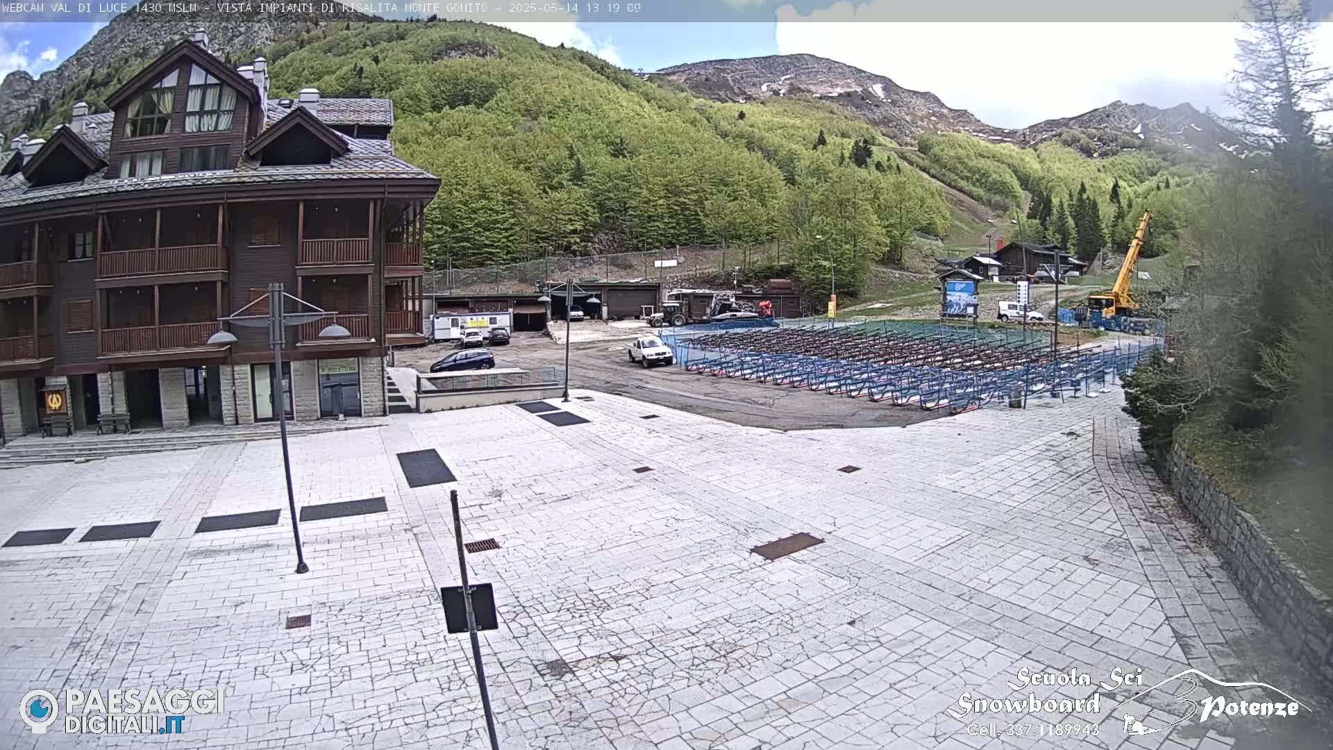 A mostly empty paved plaza sits in front of a multi-story building, with a mountainside covered in lush green trees and a partly cloudy sky in the background.