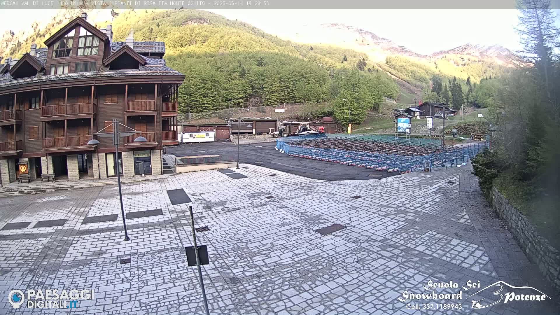 A stone plaza sits before a multi-story brown building, with a mountainside covered in green trees and some snow visible in the background under a mostly clear sky.