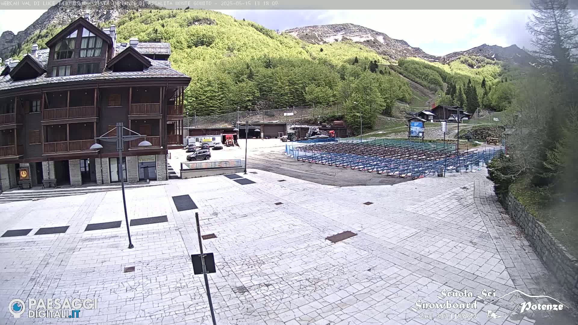 A paved plaza with a large wooden building and numerous rows of folded metal chairs sits before a mountainside covered in lush green trees, under a mostly clear sky.
