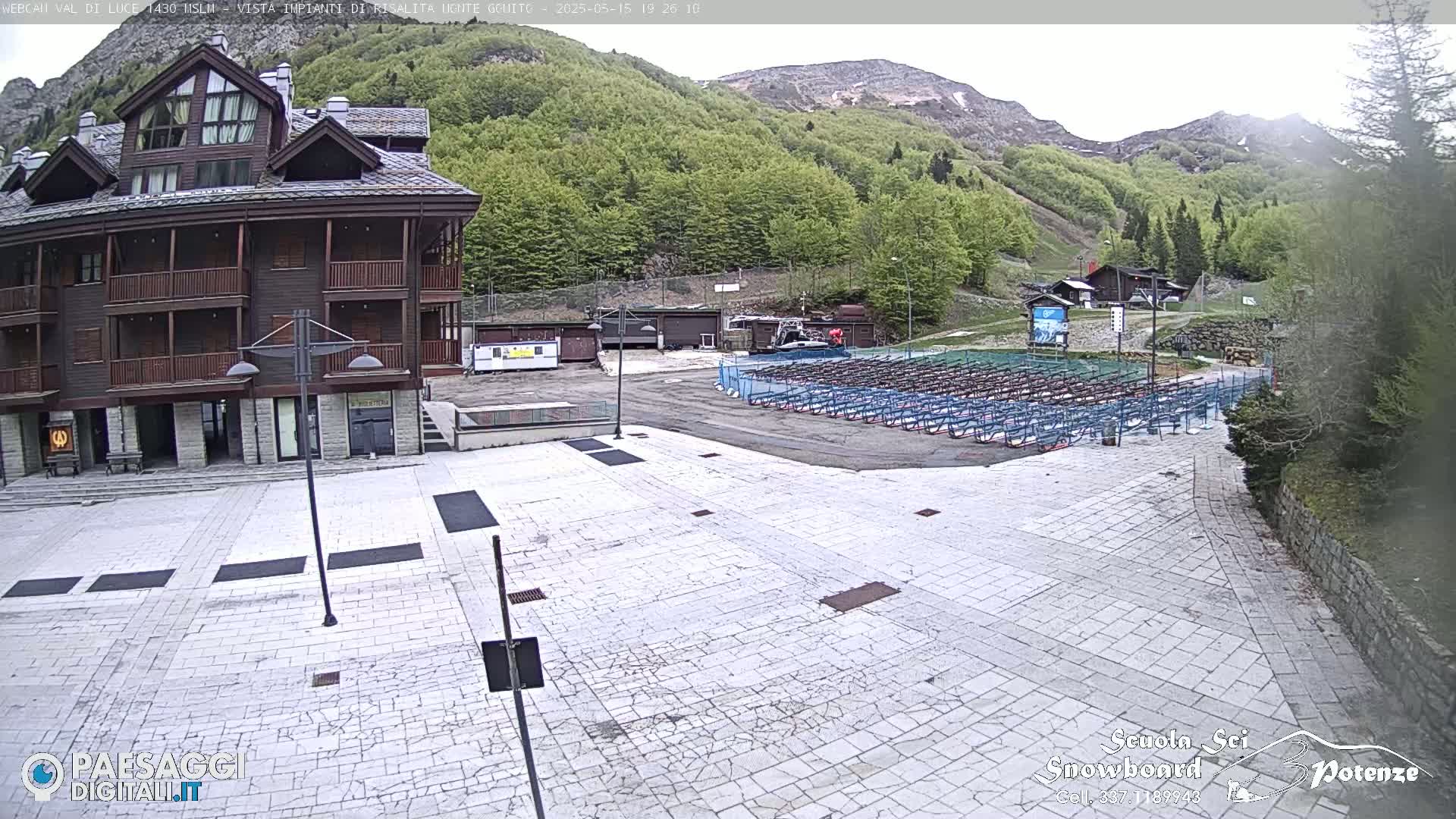 A paved plaza sits in front of a multi-story brown building, with rows of folded snowboards in the background against a backdrop of green hills under an overcast sky.