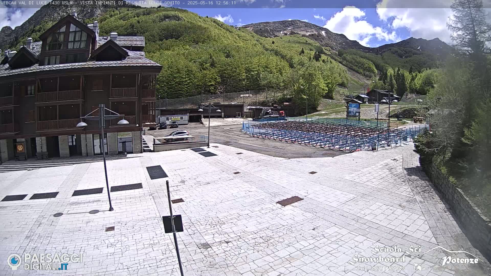 A large paved plaza sits before a multi-story brown building, with mountains and lush green trees in the background under a partly cloudy sky.