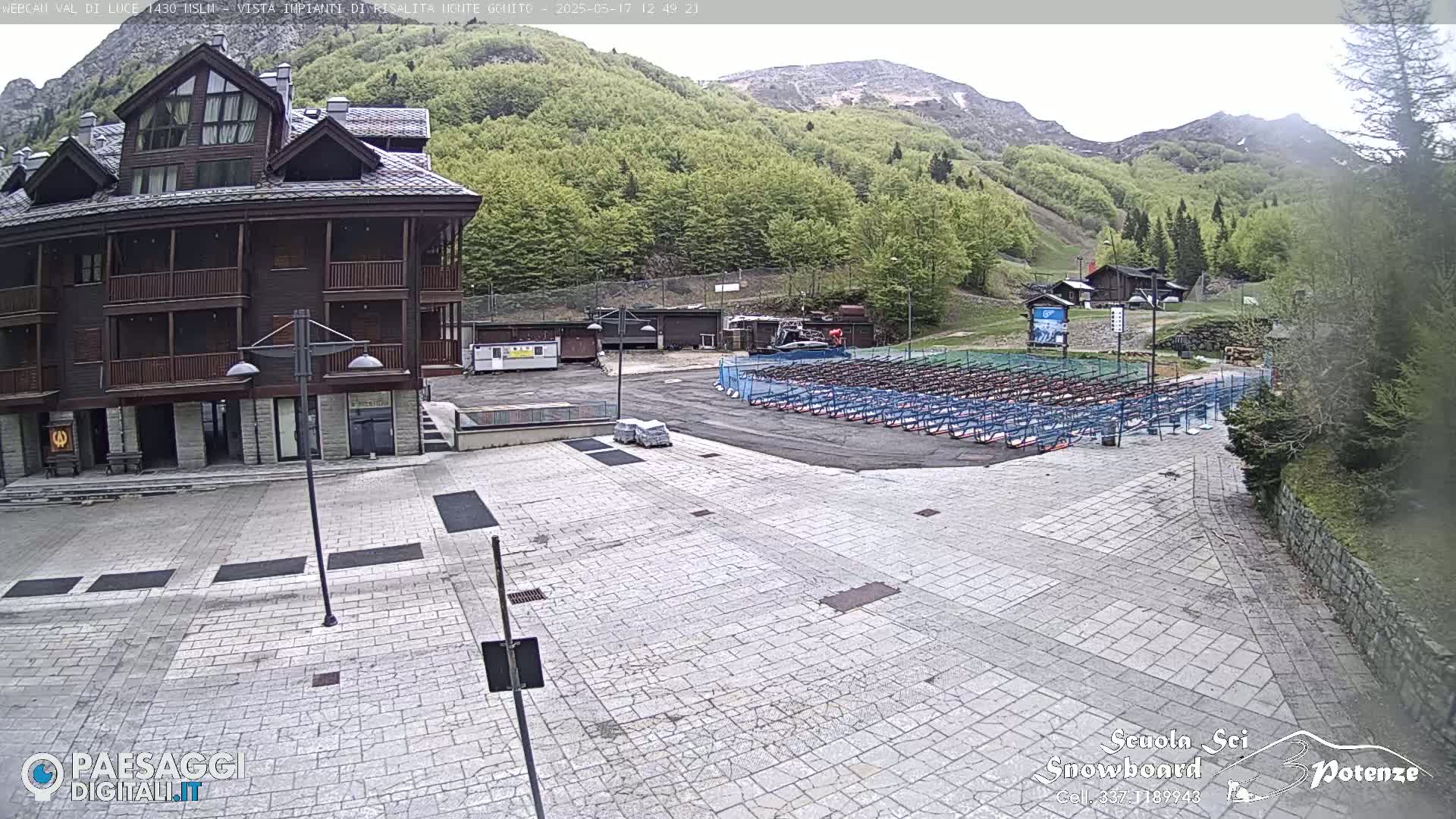 A stone plaza sits in front of a multi-story brown building, with rows of folded metal chairs or racks in the background under a partly cloudy sky, and a mountainside covered in green trees visible beyond.