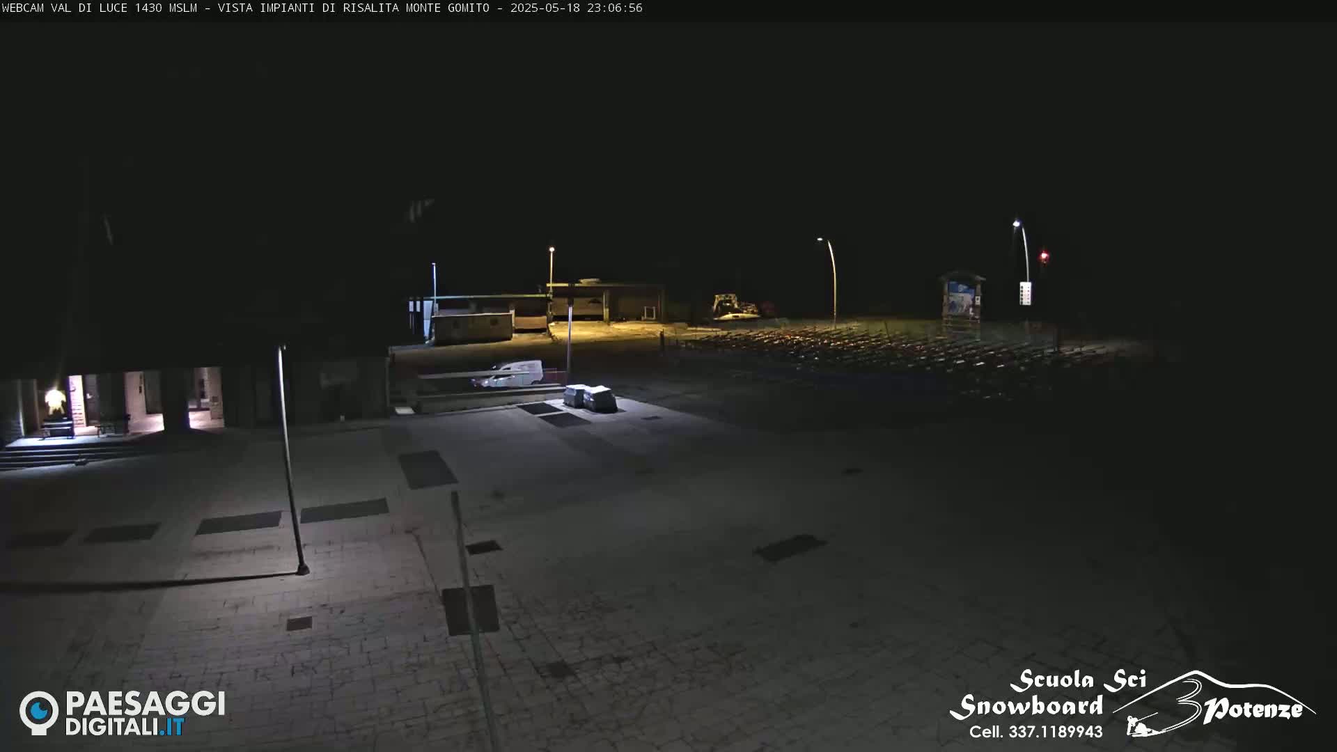 A nighttime, outdoor view of an empty paved area with some buildings and a parking lot in the background.