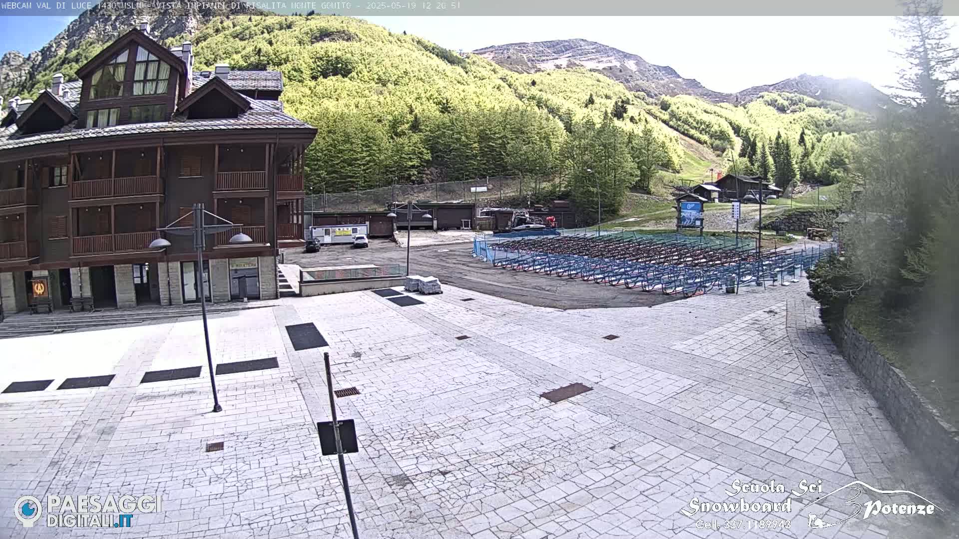 A paved plaza sits in front of a multi-story building, with a large area of folded metal racks behind it and a mountainside covered in green trees in the background under a mostly clear sky.