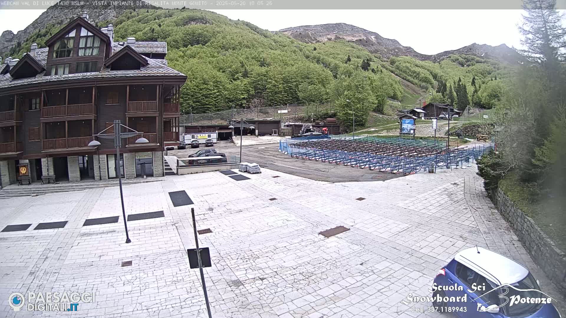 A paved plaza is in front of a multi-story wooden building, with rows of what appear to be ski or snowboard racks in the background, under a partly cloudy sky.