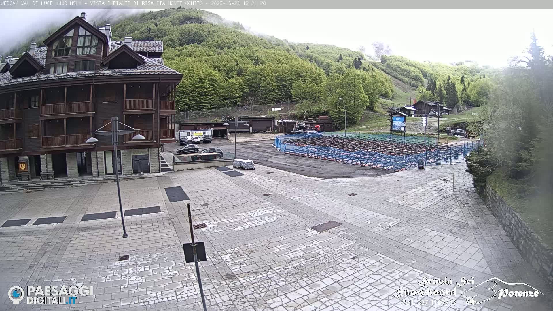 A multi-story brown building stands in a paved plaza overlooking a large area with rows of what appear to be storage racks or bike racks, all set against a background of a misty, green mountainside.