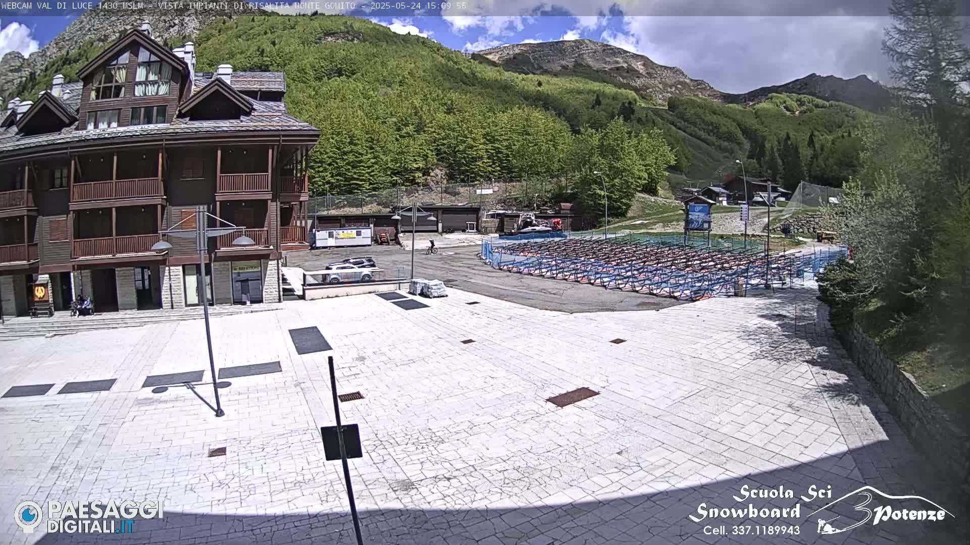 A paved plaza sits before a multi-story wood-sided building, with rows of empty equipment racks and a mountainous background under a partly sunny sky.
