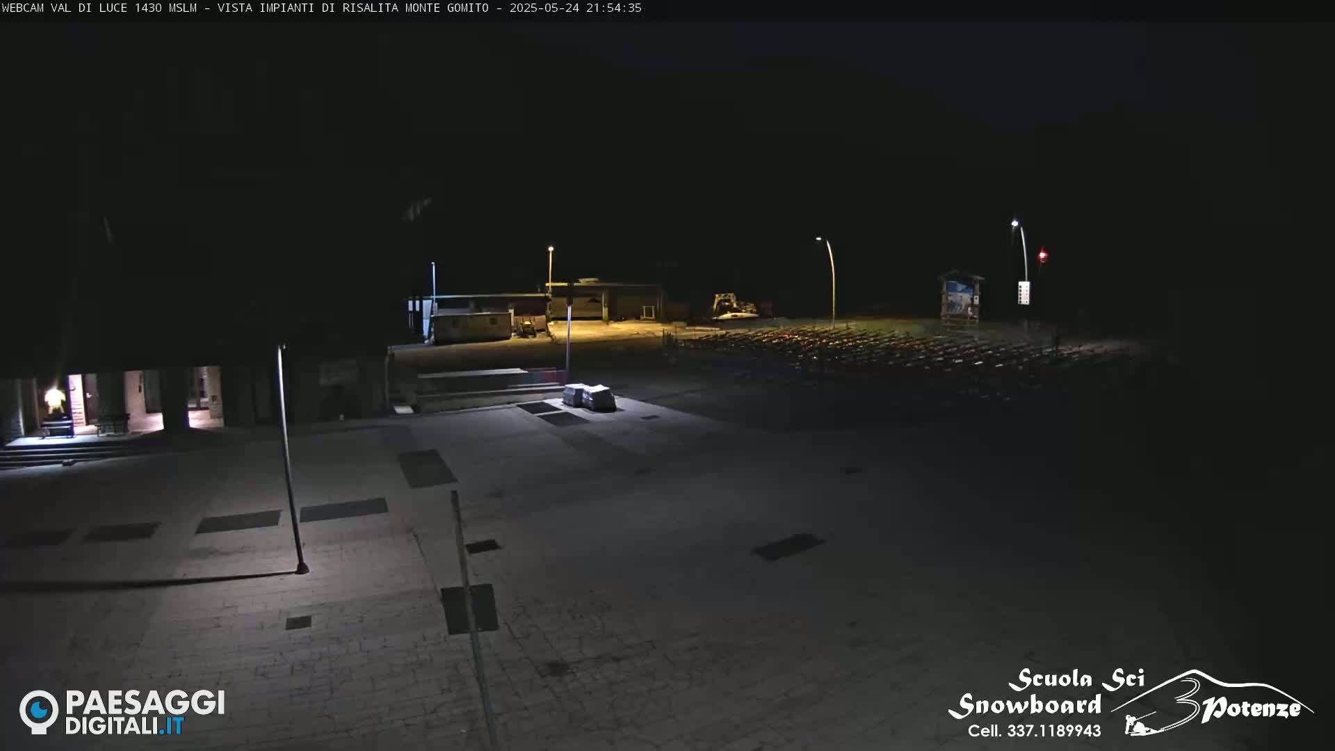 A nighttime, outdoor view of a paved plaza with buildings and a parking lot of what appear to be folded chairs or similar objects in the background.