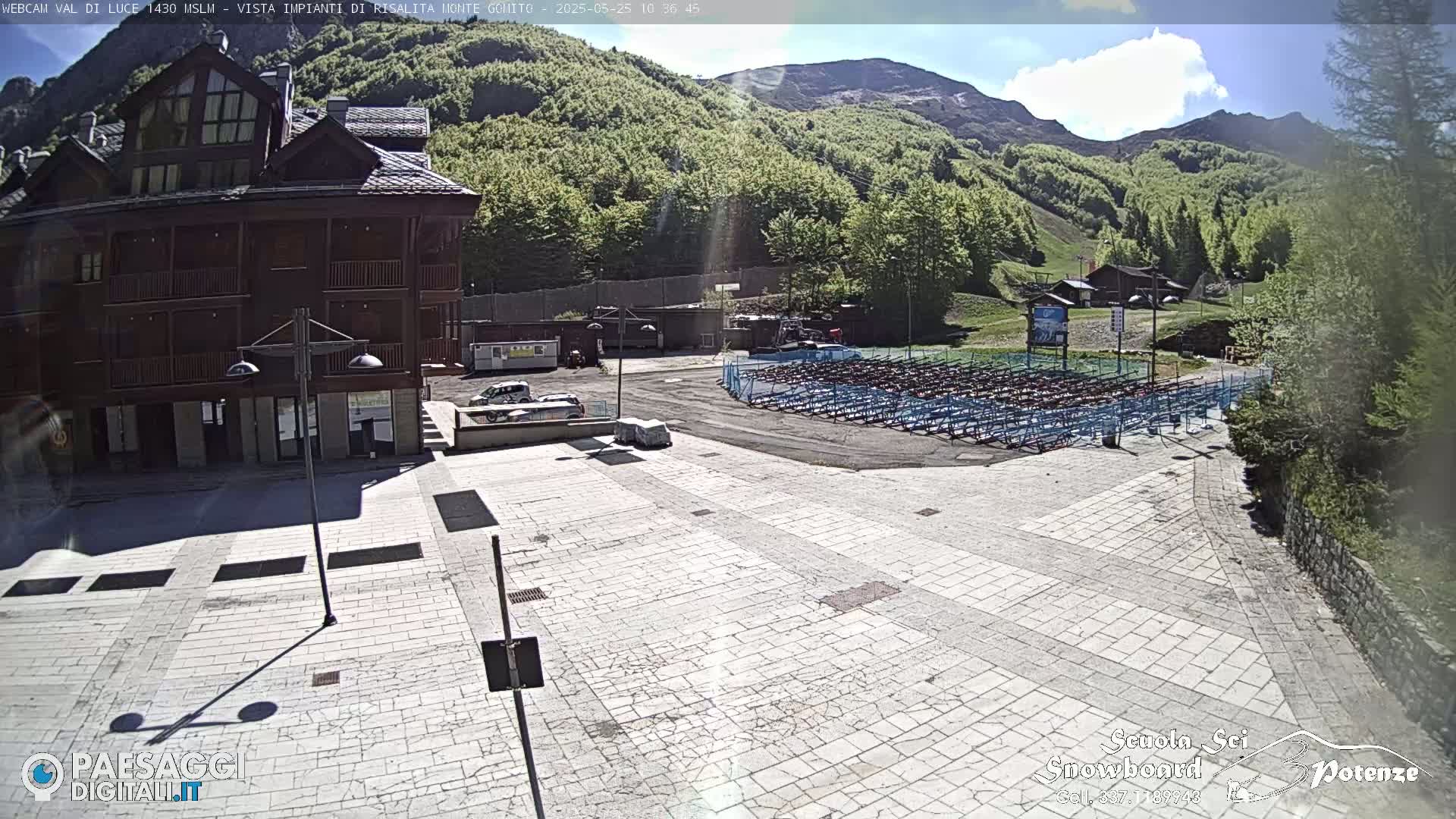 A sunny view of a plaza with a multi-story building, a mountainside with green trees, and a large area of folded chairs.